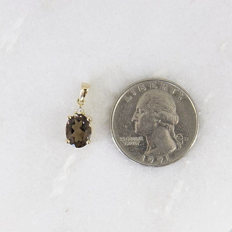 May include: A gold pendant with an oval, smoky quartz gemstone and a small diamond accent. The pendant is next to a U.S. quarter for size comparison. The gemstone is a light brown color.