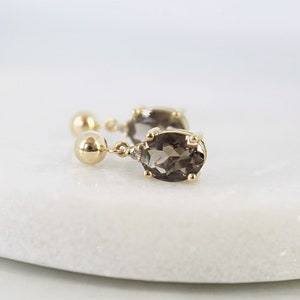 May include: A pair of gold-colored drop earrings with oval, smoky-colored gemstones. Each earring features a small, round stud, a gold chain, and a larger, oval-shaped stone. The earrings are displayed on a white surface.
