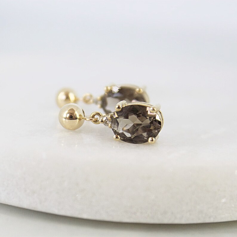 May include: A pair of gold-colored drop earrings with oval, smoky-colored gemstones. Each earring features a small, round stud, a gold chain, and a larger, oval-shaped stone. The earrings are displayed on a white surface.