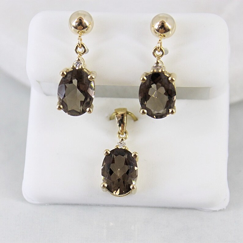 May include: A gold-toned jewelry set featuring a pair of drop earrings and a pendant. Each piece showcases an oval, smoky-brown gemstone accented with a small, clear stone. The earrings have a ball-shaped top.