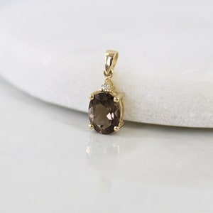 May include: A gold pendant featuring an oval, smoky quartz gemstone. The pendant has a small diamond accent and a bail for a chain. The gemstone is a rich brown color, and the setting is a polished gold.