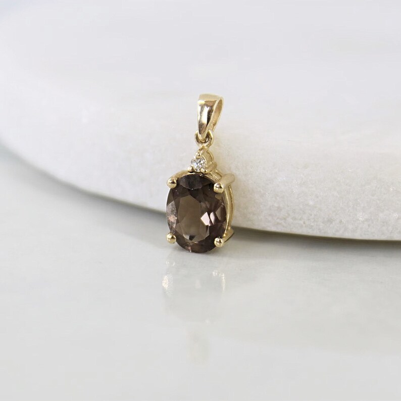 May include: A gold pendant featuring an oval, smoky quartz gemstone. The pendant has a small diamond accent and a bail for a chain. The gemstone is a rich brown color, and the setting is a polished gold.