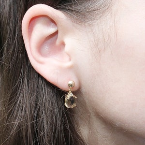 May include: Gold-toned drop earrings with a small round stud and a teardrop-shaped, light brown gemstone. The earrings are dangling near a person's ear, showcasing the jewelry's design and color.