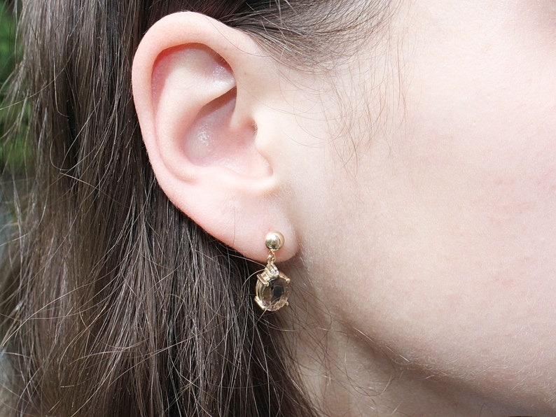 May include: Gold-toned drop earrings with a small round stud and a teardrop-shaped, light brown gemstone. The earrings are dangling near a person's ear, showcasing the jewelry's design and color.