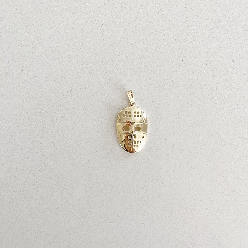 K&ouml;nnte beinhalten: Ein goldfarbener Anh&auml;nger in Form einer Hockeymaske. Die Maske hat ein detailliertes Design mit einem Netz-Muster und Augenl&ouml;chern.