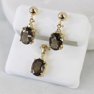 May include: A gold-toned jewelry set featuring a pendant and matching earrings. Each piece showcases a large, oval-cut, smoky quartz gemstone. The earrings dangle from a small gold ball, and the pendant hangs from a delicate chain.
