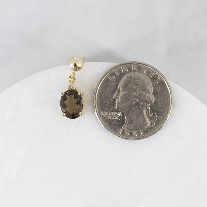 May include: A single gold earring with a smoky quartz gemstone and a small diamond accent. The oval-shaped gemstone is a deep brown color. The earring is next to a quarter for size comparison.