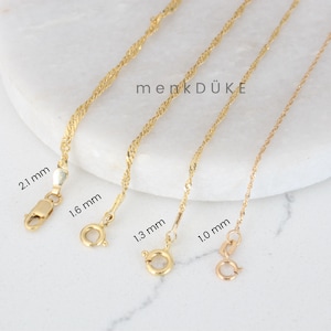 May include: Four gold-colored chain necklaces with different clasp styles and thicknesses. The chains are displayed on a white marble surface. The text "menkDÜKE" is visible in the center of the image. Measurements are provided for each chain.