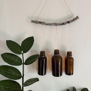 Puede incluir: Tres botellas de vidrio marrón colgando de una rama con alambre de cobre. La rama está unida a una pared blanca con alambre de cobre. Las botellas cuelgan a diferentes alturas.