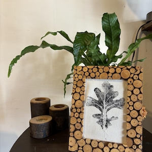 Pode incluir: Uma impressão de folha em preto e branco em uma moldura de madeira feita de pequenas peças redondas de madeira. A moldura está sobre uma mesa marrom escura com três castiçais de madeira.