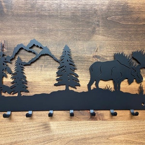 May include: Black metal moose silhouette wall hook with five hooks. The moose is standing in a forest scene with mountains and trees.