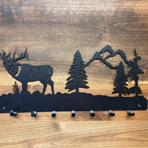 May include: Black metal key rack with a silhouette of an elk, mountains, and pine trees. The rack has six hooks.