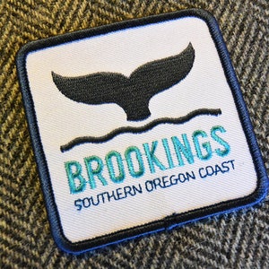 May include: Square patch with a black whale tail design above wavy lines. The text "BROOKINGS SOUTHERN OREGON COAST" is embroidered in teal and blue. The patch has a white background with a blue border.