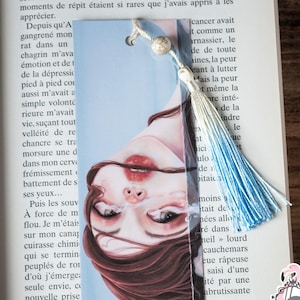 May include: A bookmark with a watercolour illustration of a woman's face, long brown hair and red lips. It has a blue and white tassel and a silver bead. The bookmark is placed in a book.