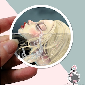 May include: Circular sticker depicting a woman's face partially submerged in water, with tears creating a splash. The woman has blonde hair, red lips, and closed eyes. The sticker has a white border and a logo in the bottom right corner.
