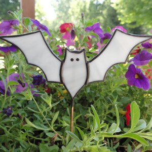 May include: A white bat-shaped garden stake with black outlines and two small black dots for eyes. The bat is set against a backdrop of colorful purple, pink, and yellow flowers and green foliage.