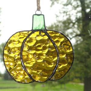 May include: Stained glass pumpkin suncatcher with a textured, yellow glass body and a green stem. The pumpkin is outlined in black and hangs from a natural twine. A decorative fall-themed item.