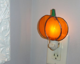 May include: A stained glass night light shaped like a pumpkin with orange glass panels and a green glass stem. The pumpkin is mounted on a white plastic plug-in base with a toggle switch, emitting a warm glow from the internal light bulb.