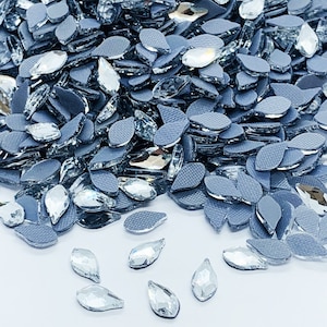 May include: A large pile of clear teardrop-shaped rhinestones with a silver backing. The rhinestones are scattered on a grey surface.