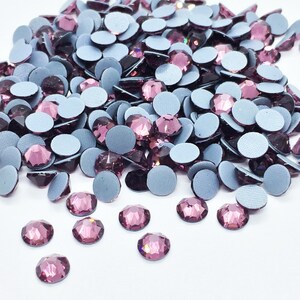 May include: A large pile of purple rhinestones with silver adhesive backs. The rhinestones are scattered on a white surface.