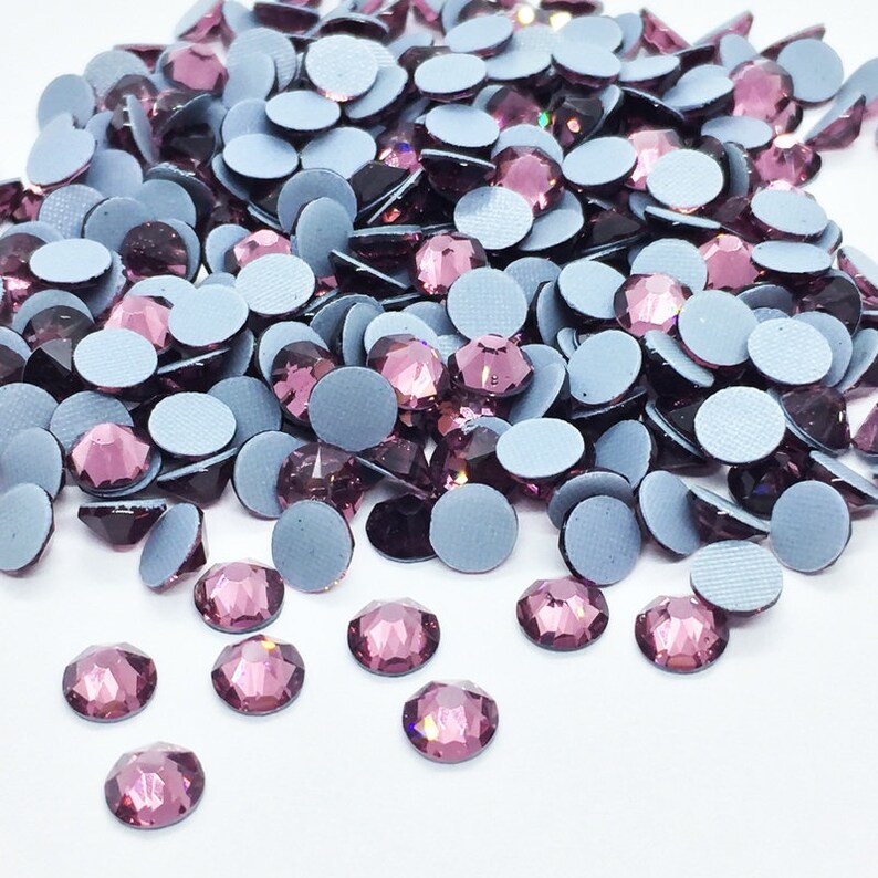 May include: A large pile of purple rhinestones with silver adhesive backs. The rhinestones are scattered on a white surface.