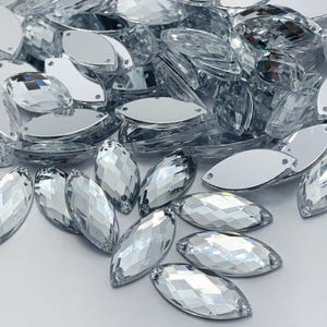 Puede incluir: Una gran cantidad de piedras de cristal transparente en forma de lágrima. Las piedras tienen un diseño facetado y están esparcidas sobre una superficie blanca.
