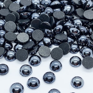 Könnte beinhalten: Ein großer Haufen schwarzer, runder, flach geformter, irisierender Glascabochons. Die Cabochons sind auf einer weißen Oberfläche verstreut.