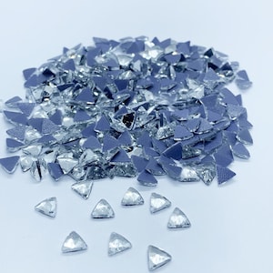 Puede incluir: Un montón grande de piedras de cristal triangulares transparentes con un respaldo plateado. Las piedras de cristal están esparcidas sobre una superficie blanca, con algunas piedras de cristal más pequeñas esparcidas alrededor del montón.