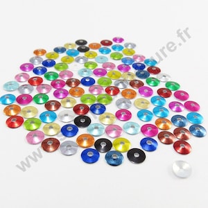 Può includere: Assortimento di paillettes rotonde metalliche colorate con un centro rialzato. Le paillettes sono sparse su una superficie bianca.
