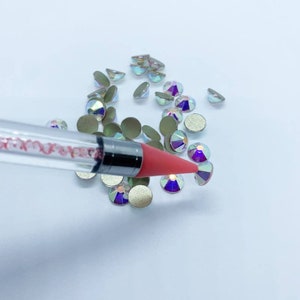 Op de afbeelding: Een close-up van een transparant plastic gereedschap met een rode punt, gebruikt voor het aanbrengen van strass steentjes. Het gereedschap ligt op een wit oppervlak met een strooiing van iriserende strass steentjes.