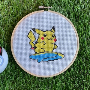 Puede incluir: Un patrón de punto de cruz Pikachu amarillo en una tela blanca estirada sobre un aro de bordado de madera. El Pokémon está montando una tabla de surf azul.