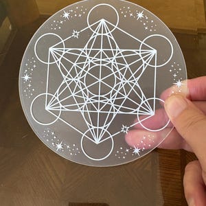 May include: A clear, round crystal grid with a white Metatron's Cube design. The design features geometric shapes and star accents around the edge. The crystal grid is held by a hand.
