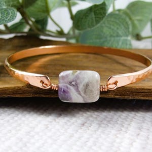 May include: A copper-coloured bangle bracelet with a hammered texture. The bracelet features a square, light purple and white stone at the centre. The bracelet is displayed on a wooden surface.