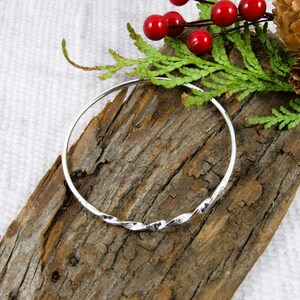 May include: A silver bangle bracelet with a twisted design. The bracelet is displayed on a piece of wood, decorated with red berries, green sprigs, and a pine cone. The background is a white, textured surface.