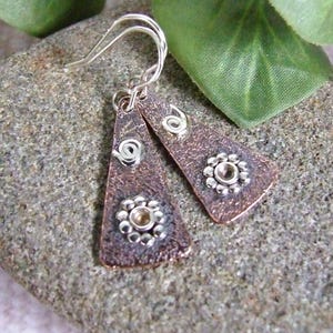 May include: Copper-toned earrings with silver detailing. Each earring features a textured, triangular shape with a silver spiral and a flower design. The earrings are suspended from silver hooks.