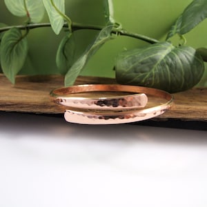 May include: A copper-coloured, hammered-finish bracelet with a spiral design. The bracelet wraps around the wrist with an open end. The metal has a warm, reddish-brown hue. The bracelet is displayed on a wooden surface with green foliage in the background.