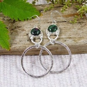 May include: Silver hoop earrings with a textured finish. Each earring has a dark green gemstone in a decorative silver setting. The earrings are displayed on a piece of wood. The hoops are approximately 4 cm in diameter.