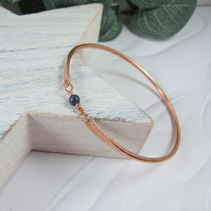 May include: A rose gold bangle bracelet with a single dark blue bead accent.