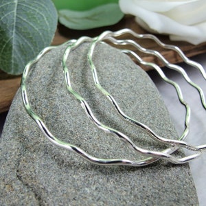 May include: Three silver bangle bracelets with a wavy design. The bracelets are stacked on top of each other.