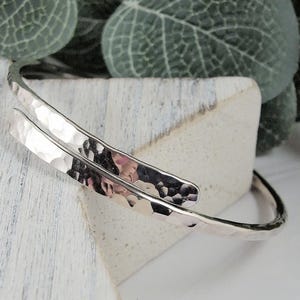 May include: A silver hammered bangle bracelet with a unique double-wrap design. The bracelet is made from a single piece of metal that is bent and hammered to create a textured surface.