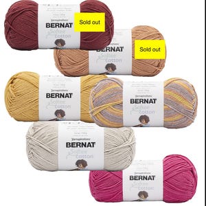 May include: A collection of Bernat Softee Cotton yarn skeins in various colors, including burgundy, tan, yellow, and pink. Some skeins have "Sold out" labels. Each skein is labeled with the brand name and product details.