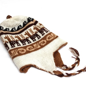 May include: A knitted hat with ear flaps and a braided tassel. The hat features a pattern of brown llamas on a white background, with brown and black geometric designs. The hat is made of a soft, warm material.