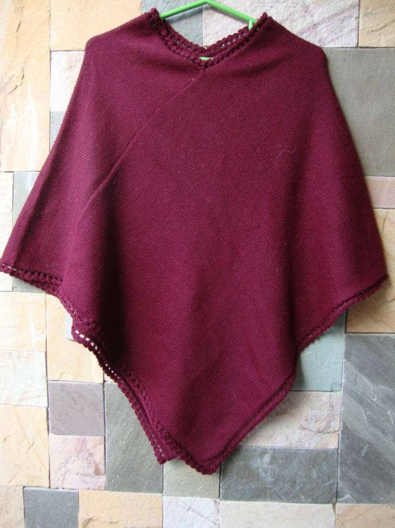 Pode incluir: Um poncho de malha bord&ocirc; com uma borda de croch&ecirc;. O poncho est&aacute; pendurado em um cabide contra uma parede de azulejos.