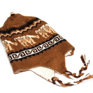 May include: A brown knit hat with ear flaps and a braided tassel. The hat features a repeating pattern of white and tan llamas and geometric designs. The hat is made of a soft, warm material.