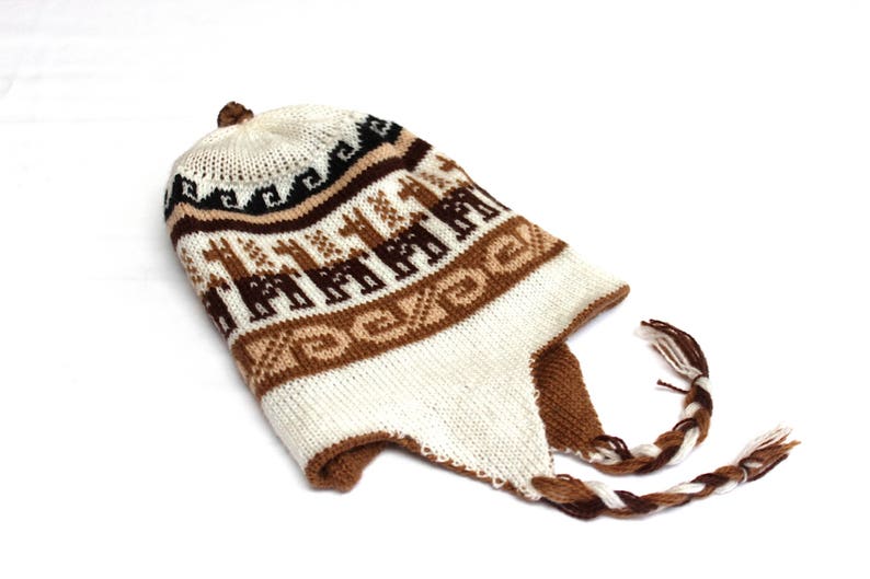 May include: A knitted hat with ear flaps and a braided tassel. The hat features a pattern of brown llamas on a white background, with brown and black geometric designs. The hat is made of a soft, warm material.
