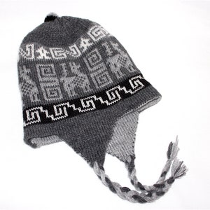 May include: Gray knit hat with ear flaps and a braided tassel. The hat features a geometric pattern in white and black, with the word "JAG" visible. A small black pom-pom sits on top.