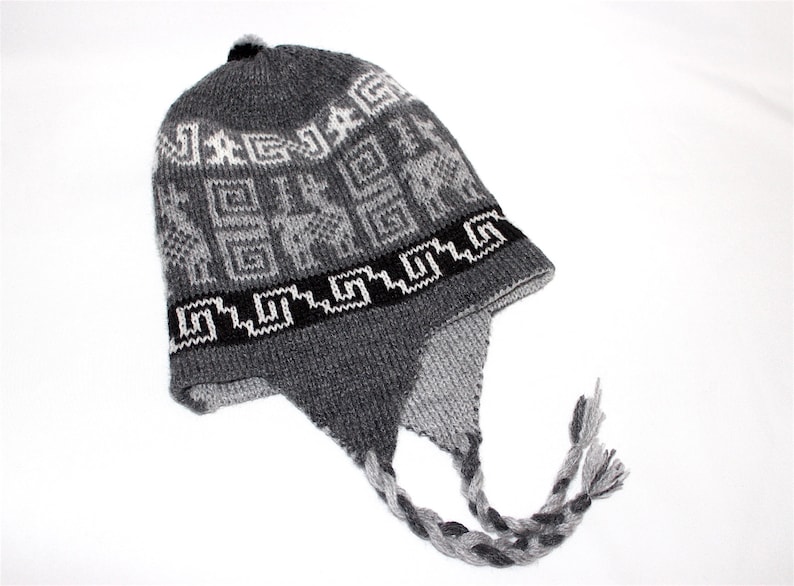 May include: Gray knit hat with ear flaps and a braided tassel. The hat features a geometric pattern in white and black, with the word "JAG" visible. A small black pom-pom sits on top.