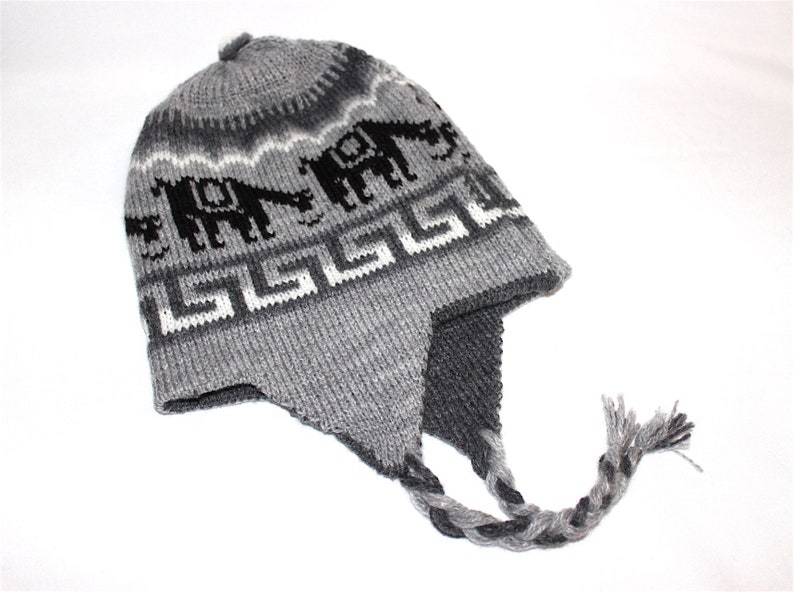 May include: Gray knit hat with ear flaps and a braided tassel. The hat features a repeating pattern of black elephants and white geometric shapes against a gray background. A cozy winter accessory.