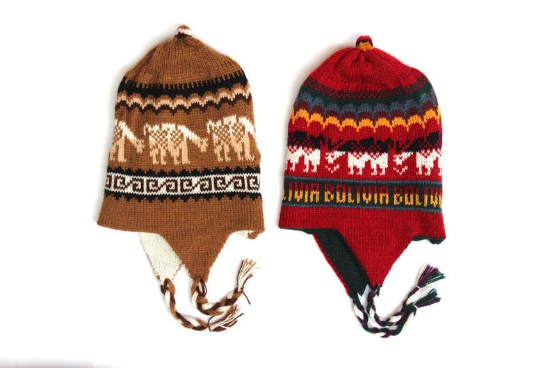 May include: Two knitted alpaca hats. One is brown with llama designs, the other is red with a geometric pattern and the text "Bolivia". Both have ear flaps and tassels. These hats are perfect for cold weather.