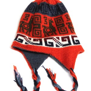 May include: A knitted hat with ear flaps, featuring a vibrant red base with geometric patterns in dark blue and brown. The hat has a braided tassel and a small orange pom-pom on top. The ear flaps are dark blue and the tassels are a mix of blue, orange, and grey.